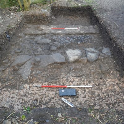 Trench 11 showing earlier phase stone wall foundation (foreground) with the later wall of the 18th century mansion house in the background. A rough cobbled floor surface lies in between. © Copyright ARS Ltd 2018